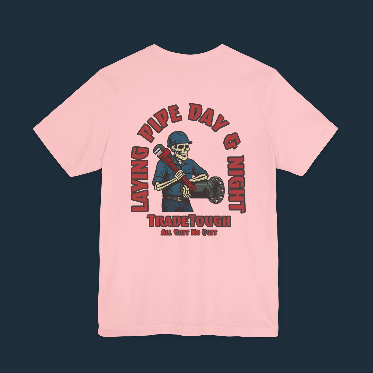 Pink t-shirt with a graphic design on the back featuring a skeleton wearing a hard hat and holding a pipe, with text 'Laying Pipe Day & Night' and 'Tradetough'.