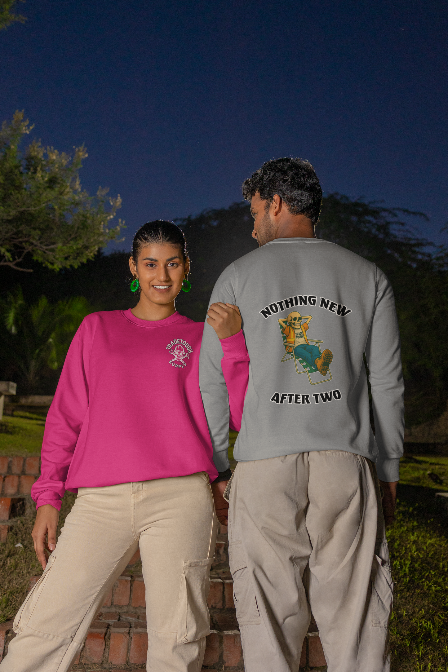 Two people walking outdoors at night, one wearing a pink sweatshirt with a logo and the other wearing a gray sweatshirt with a graphic design.